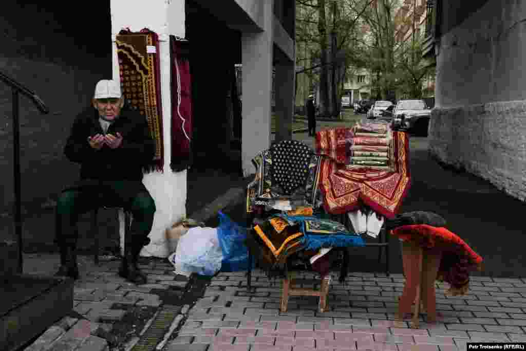 Орталық мешіттің маңында жайнамаз бен басқа да заттар сатып отырған саудагер намаз оқылып жатқан кезде қол жайып, дұға қылып отыр.